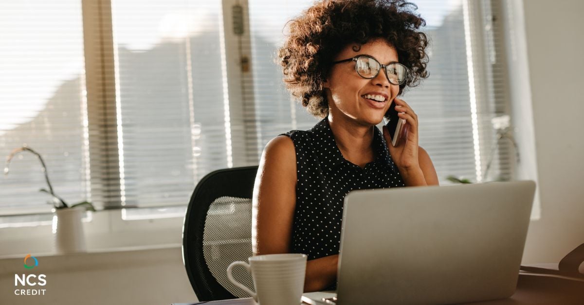 woman at desk on cell phone in front of laptop