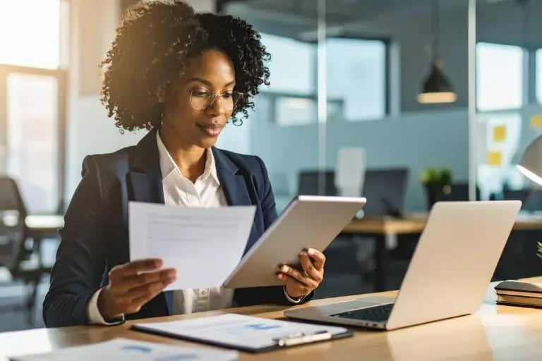 grinning business woman looking at tablet and document at desk with laptop and clipboard