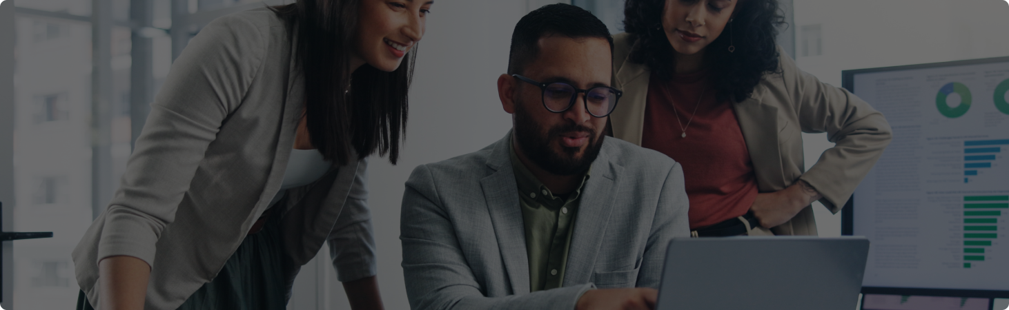 two business woman standing at a business man's desk looking over his shoulder at his laptop