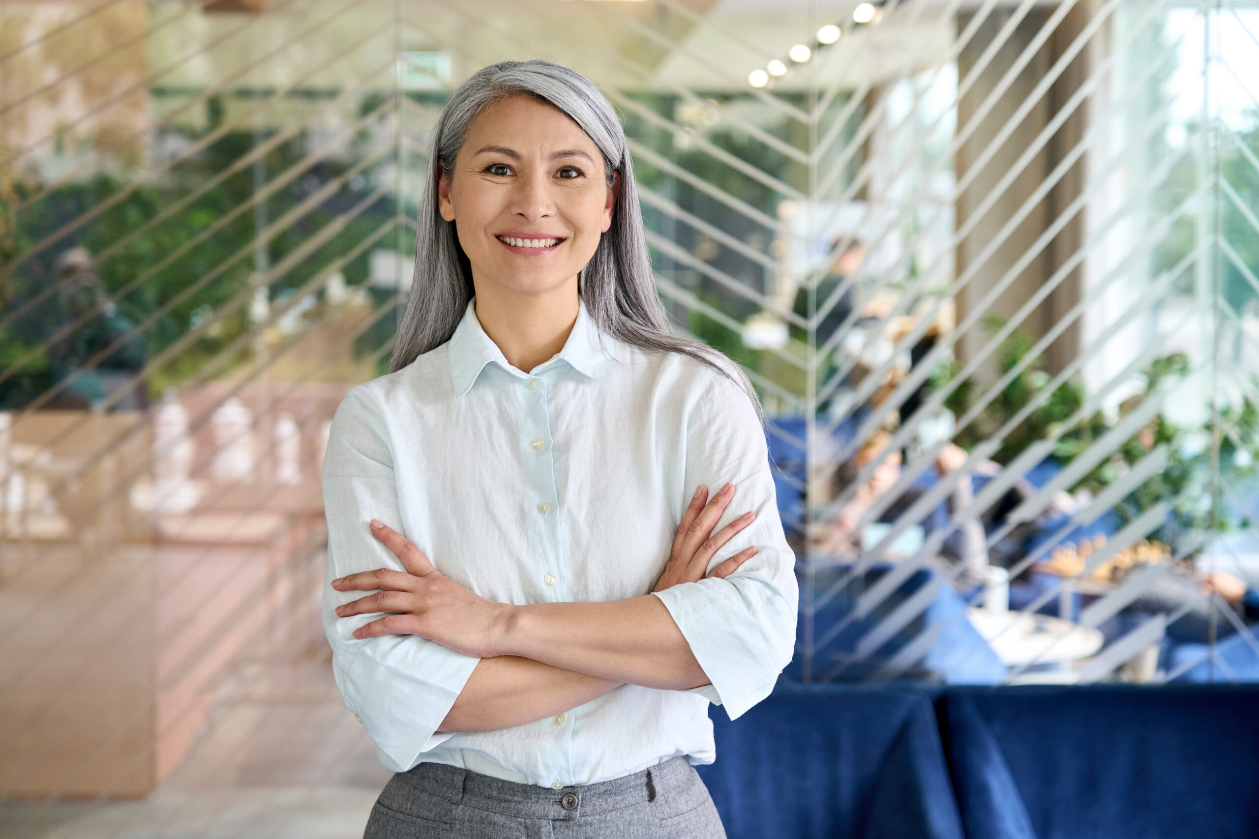 Happy smiling confident middle aged Asian older senior female leader businesswoman standing in modern office workplace looking at camera arms crossed. Business successful executive concept. Portrait.